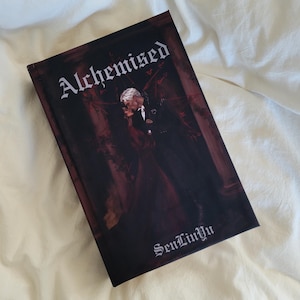 May include: A hardcover book titled Alchemised by SenLinYu with a dark, moody cover illustration featuring two figures in formal attire embracing in a dimly lit, red-toned setting with architectural columns and intricate, dark floral details.