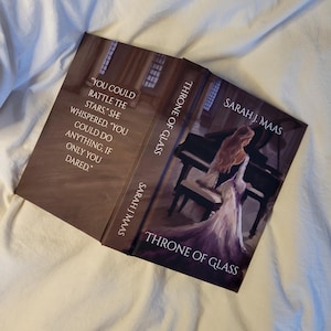 Peut inclure: Un livre ouvert intitulé "Throne of Glass" de Sarah J. Maas. La couverture représente une femme en robe blanche près d'un piano. La couverture du livre comporte une citation : "You could rattle the stars..." sur le côté gauche.