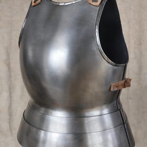 May include: A steel breastplate with leather straps and buckles. The armour has a curved shape and a layered lower section. The metal has a reflective surface, and the leather straps are a light brown colour.