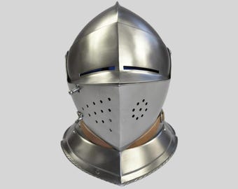 Puede incluir: Un casco medieval pulido de color plateado. El casco presenta una parte superior puntiaguda, una placa frontal con orificios de ventilación y un protector de cuello. El diseño incluye remaches y una correa de cuero, lo que sugiere artesanía histórica. El casco es un símbolo de protección.