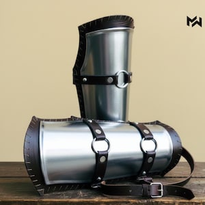 Puede incluir: Dos piezas de armadura medieval para el brazo. Los protectores de brazo metálicos son cilíndricos con ribetes y correas de cuero marrón. Cada pieza tiene un anillo metálico y una hebilla para asegurar la armadura. El fondo es de color beige claro.