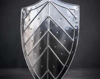 Pode incluir: Um escudo de metal prateado polido com formato pontiagudo, apresentando uma crista central vertical e linhas em relevo simétricas com padrão de chevron. A superfície é acentuada com pequenos rebites espaçados uniformemente ao longo das bordas.