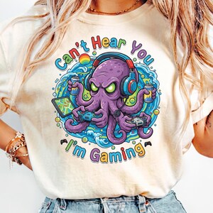 Puede incluir: Camiseta color crema con un pulpo morado con auriculares y sosteniendo mandos de juego y bebidas. El pulpo está rodeado de agua y el texto "Can't Hear You I'm Gaming" en un diseño colorido.