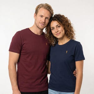 Puede incluir: Dos personas con camisetas a juego. Una es de color burdeos, la otra azul marino. Ambas tienen un pequeño diseño dorado bordado en el pecho izquierdo. Las camisetas de manga corta parecen estar hechas de un material suave.