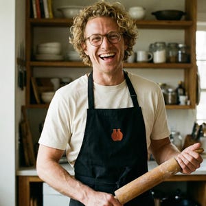 May include: A smiling person wearing glasses and a black apron with a brown emblem, holding a wooden rolling pin. The person is in a kitchen with wooden shelves filled with kitchenware.