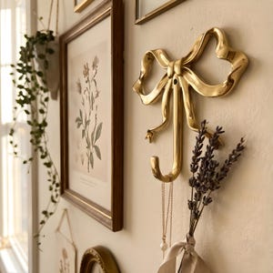 Może przedstawiać: Złoty metalowy wieszak ścienny w kształcie kokardy. Z wieszaka zwisa bukiet lawendy i naszyjnik. Wieszak jest zamontowany na kremowej ścianie, z oprawionymi botanicznymi nadrukami i innymi widocznymi przedmiotami.