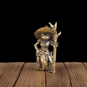 May include: A detailed brass figurine of a frog dressed in fantasy attire, including a wide-brimmed hat and a staff. The frog is posed with one hand on its hip and the other holding the staff. The figurine has intricate details and a vintage aesthetic.