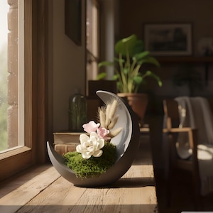 May include: A crescent moon-shaped decorative planter in a dark gray color. The planter holds green moss, white and pink flowers, and dried wheat. The planter sits on a wooden surface near a window.