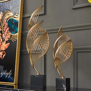 Puede incluir: Dos esculturas de metal dorado con un diseño en forma de hoja. Cada escultura está compuesta por líneas curvas e entrelazadas y se asienta sobre una base cúbica de mármol negro. Son objetos de arte decorativos.