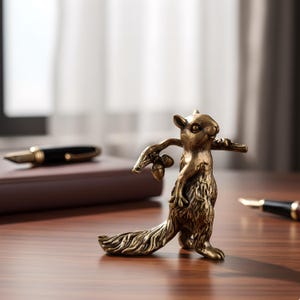 May include: A bronze squirrel figurine with outstretched arms holding acorns. The detailed sculpture has a textured finish and stands on a wooden surface. A pen and notebook are in the background.