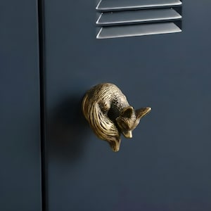 Pode incluir: Um puxador de armário em forma de raposa de latão montado numa porta de armário azul escuro. A raposa está enrolada, com textura de pelo detalhada. O puxador adiciona um toque de decoração inspirada na natureza.
