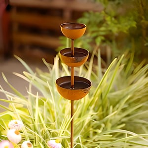 May include: A tiered, outdoor metal bird feeder with three bowl-shaped levels. The feeder has a rusty orange finish and is set against a backdrop of green foliage. The bowls are designed to hold birdseed or water, attracting birds to a garden.