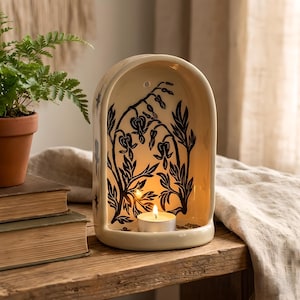 Könnte beinhalten: Cremefarbener Keramik-Kerzenhalter mit schwarzem Blumendesign. Der bogenförmige Halter enthält eine brennende Teelichtkerze, die einen warmen Schein erzeugt. Der Halter steht auf einer Holzoberfläche, neben einer Topffarn und alten Büchern.