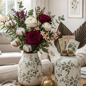 Puede incluir: Dos jarrones de cerámica blanca con motivos florales verdes sostienen un vibrante arreglo floral. El arreglo incluye tallos de algodón, ranúnculos blancos y rosas rojo oscuro. Un pequeño pulverizador de latón y herramientas de jardinería también están sobre la mesa de madera.