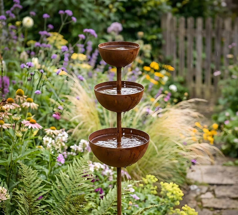 May include: A tiered outdoor water fountain with three bowl-shaped basins. The fountain is made of rusty brown metal and is set against a backdrop of a lush garden with colorful flowers and greenery. Water flows from the top basin to the bottom.