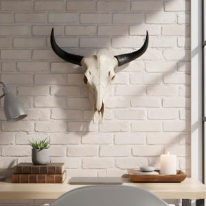 May include: A cow skull with black horns is mounted on a white brick wall. The skull is cream-coloured. A desk with a laptop, books, a candle, and a small plant is in front of the wall.