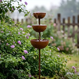 May include: A three-tiered outdoor water fountain made of weathered metal. Water cascades from the top bowl to the bottom, creating a tranquil effect. The fountain is set in a garden with green foliage and purple flowers, with a wooden fence in the background.