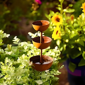 May include: A tiered, rustic-style metal bird bath with three bowl-shaped levels. The bird bath is a warm, rusty brown color and is set against a backdrop of green foliage and yellow flowers.