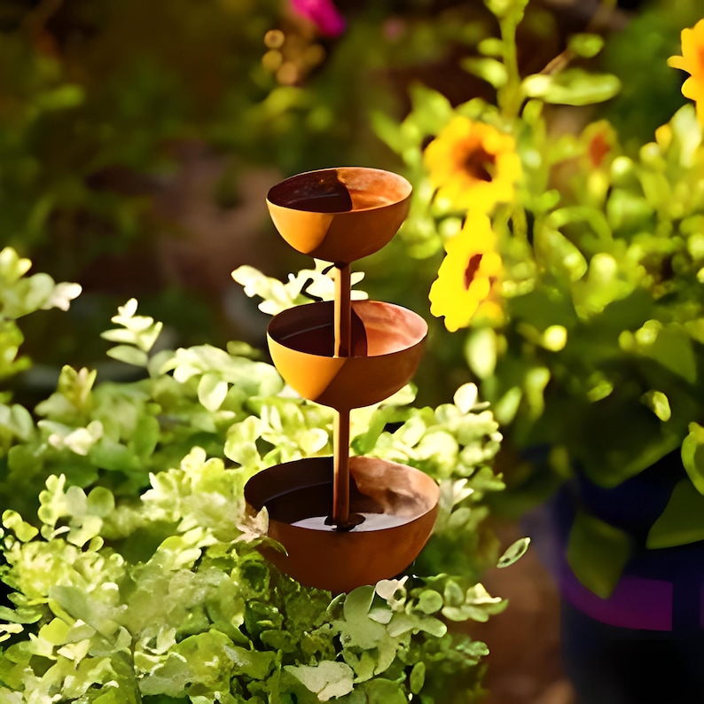 May include: A tiered, rustic-style metal bird bath with three bowl-shaped levels. The bird bath is a warm, rusty brown color and is set against a backdrop of green foliage and yellow flowers.