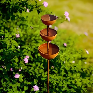 May include: A three-tiered, bronze-colored bird feeder with a slender pole. Each tier is a shallow bowl, perfect for holding birdseed. The feeder stands in front of a backdrop of green foliage and small pink flowers.