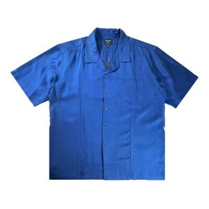 Può includere: Una camicia a maniche corte con abbottonatura in un colore blu reale tinta unita. La camicia presenta un colletto a revers, una tasca singola sul petto sul lato sinistro e una cucitura verticale sui pannelli frontali. Un'etichetta scura al collo.