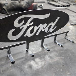 May include: A metal wall-mounted coat rack featuring the Ford logo. The logo is cut out of a dark oval shape, with the word "Ford" in a lighter color. The rack has four hooks below the logo.