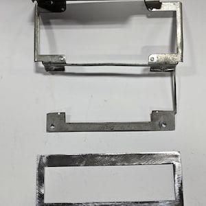 May include: A collection of three metal components, likely for a mechanical device. The pieces are silver-toned and rectangular, with various mounting holes and a frame-like structure. The metal has a brushed appearance, suggesting a utilitarian design.