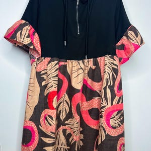 Könnte beinhalten: Schwarzes Kapuzenkleid mit Reißverschluss. Die Ärmel haben ein rosa-braunes Blättermuster. Der untere Teil des Kleides hat einen braunen Hintergrund mit rosa, roten und beigen Blatt- und schlangenartigen Mustern.