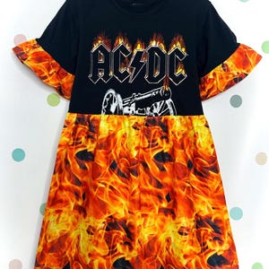 Puede incluir: Vestido negro con el logo de AC/DC en blanco y plata. La parte superior es negra, y la falda y los bordes de las mangas tienen un diseño de llamas naranja y amarillo vibrante. El vestido tiene cuello redondo y mangas cortas. Estilo rock.
