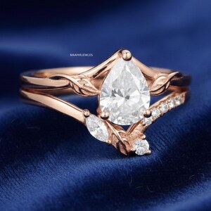 Pode incluir: Conjunto de anéis de noivado em ouro rosa com uma pedra central de diamante em forma de pêra. O anel apresenta um design de folha e uma banda com diamantes menores. O anel está sobre um fundo azul.
