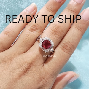 Puede incluir: Un anillo de tono plateado con una piedra preciosa central de color rojo intenso en talla cojín, rodeada por piedras blancas transparentes más pequeñas en un engaste de halo. El texto READY TO SHIP aparece arriba y MAAHIRJEWEL debajo.