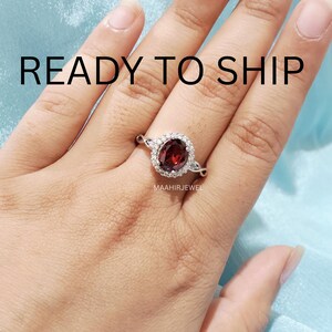 May include: A silver ring featuring a central oval-cut deep red garnet gemstone surrounded by a halo of small white stones. The band has a twisted design and the text READY TO SHIP and MAAHIRJEWEL are visible on the image surface.