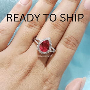 Könnte beinhalten: Ein silberfarbener Ring mit einem birnenförmigen roten Mittelstein, der von einem Kranz aus kleinen klaren Steinen umgeben ist. Die Ringschiene ist geteilt und mit winzigen klaren Steinen besetzt. Der Text READY TO SHIP und MAAHIRJEWEL ist sichtbar.