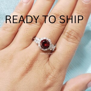 May include: A silver ring featuring a round, deep red garnet center stone surrounded by a halo of small, clear white accent stones. The text READY TO SHIP is visible at the top, and the text MAAHIRJEWEL is printed below the ring setting.