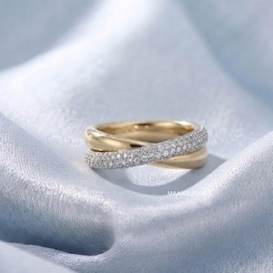 May include: A crossover ring featuring two bands, one in polished yellow gold and the other encrusted with small white pavé stones. The ring displays the text MAAHIRJEWELS in small white letters on the lower right side of the band structure.