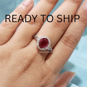 May include: A silver-toned ring featuring a central oval-cut deep red gemstone surrounded by a halo of small clear stones. The band has a twisted design. The text READY TO SHIP is visible at the top and MAAHIRJEWEL is printed below the ring.