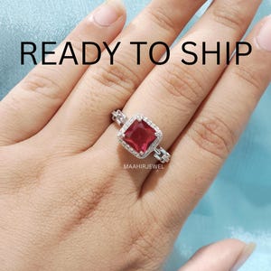 Könnte beinhalten: Ein silberfarbener Ring mit einem quadratisch geschliffenen, tiefroten Mittelstein, der von einem Kranz aus kleinen, klaren Steinen umgeben ist. Die Ringschiene hat ein dekoratives Gliederkettendesign. Text READY TO SHIP und MAAHIRJEWEL sichtbar.