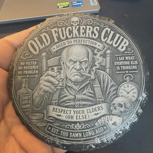 Op de afbeelding: Een ronde, donkergrijze onderzetter met een gedetailleerde illustratie van een oude man die een sigaar rookt en drinkt. Tekst bevat "Old Fuckers Club" en "Respect Your Elders (Or Else)".