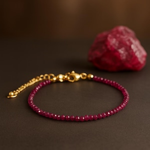Op de afbeelding: Een delicate armband met kleine, gefacetteerde robijnkralen. De armband heeft een goudkleurige sluiting en ketting. Een ruwe, rode robijnsteen staat op de achtergrond. Sieraden.