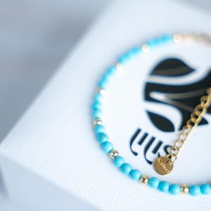 Könnte beinhalten: Ein türkisfarbenes Perlenarmband mit goldenen Akzenten und einer goldenen Kette. Das Armband liegt auf einer weißen Schachtel mit einem schwarzen Logo. Ein kleiner goldener Anhänger trägt die Aufschrift "s.steel".