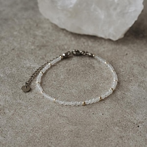 Op de afbeelding: Delicate armband met kleine, doorschijnende kralen en goudkleurige accenten. De armband heeft een zilverkleurige ketting en sluiting. Een witte, getextureerde steen staat op de achtergrond. Sieraden.