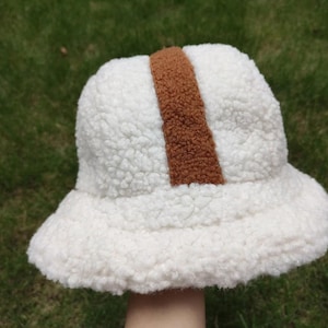 May include: A white, fluffy bucket hat with a brown vertical stripe. The hat is made of a soft, textured material, and the brim is slightly curved. This cozy hat is perfect for adding warmth and style to any outfit.