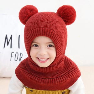 May include: A red knitted hat and cowl set for children. The hat features two fluffy pom-poms on top and a built-in cowl that covers the neck and shoulders. The text "MI FOR" is visible in the background.