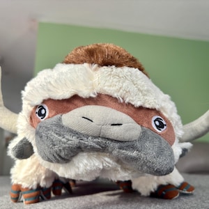 May include: A plush toy of a bison-like creature with white fur, brown accents, and large, expressive eyes. The toy has large, curved horns and is posed on a gray surface. The creature's feet have striped details.