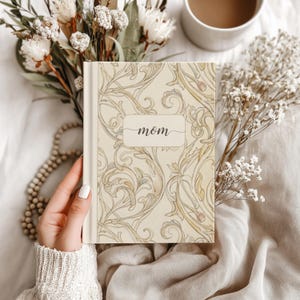 Może przedstawiać: Twarda oprawa dziennika w kremowym kolorze, z wzorem kwiatowym w kolorze złota i zieleni. Słowo "mom" jest napisane kursywą na etykiecie. Dziennik otoczony jest kwiatami i filiżanką kawy, tworząc spokojną estetykę.