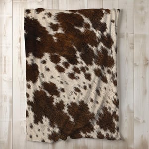 May include: A rectangular throw blanket featuring a realistic brown and white cowhide print. The pattern consists of irregular, organic dark brown patches distributed across a creamy white background, creating a classic spotted animal hide aesthetic.