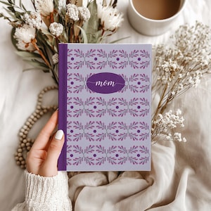 May include: A purple journal with the word "Mom" in cursive on the cover. The cover has a light purple background with a repeating floral design in purple and pink. The journal is held by a person.