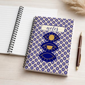 Puede incluir: Cuaderno de espiral con un patrón de ojos azules y dorados y la palabra "notes" en la portada. También se ven un cuaderno abierto y un bolígrafo.
