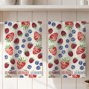 Puede incluir: Dos paños de cocina con un estampado de mosaico de fresas rojas, frambuesas y arándanos azules sobre un fondo crema. Los paños tienen el texto francés "Les Framboises, Les Fraises, Les Bleuets" en la parte inferior.