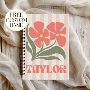 Puede incluir: Un cuaderno de espiral con un fondo de rayas verticales en rosa claro y blanco. La portada muestra dos flores grandes de color coral con centros amarillos y hojas verde salvia. El nombre TAYLOR está impreso en color coral abajo.